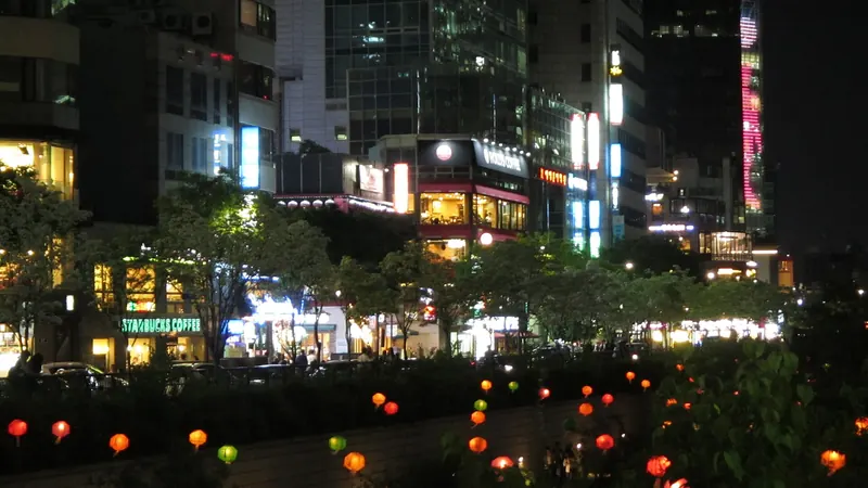 Glowing red and green lanterns along the Cheonggyecheon stream at night, below lit office towers in central Seoul