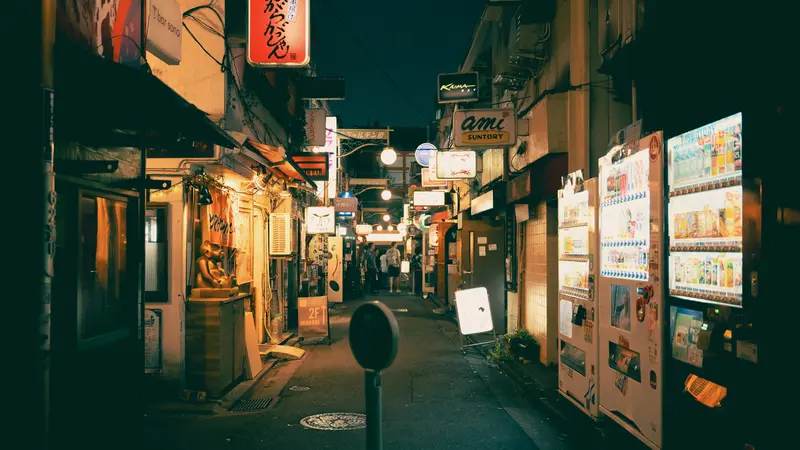 A narrow alley in Shinjuku at night with glowing shop signs and a row of lit vending machines