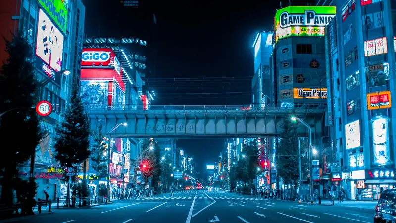 Rua vazia em Akihabara, Tóquio, à noite, iluminada por letreiros azuis e de neon, com uma linha férrea elevada cruzando a via