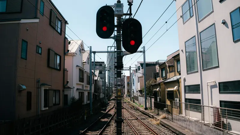Railway tracks running between apartment buildings in a Tokyo neighborhood, with red signal lights and overhead wires under a blue sky