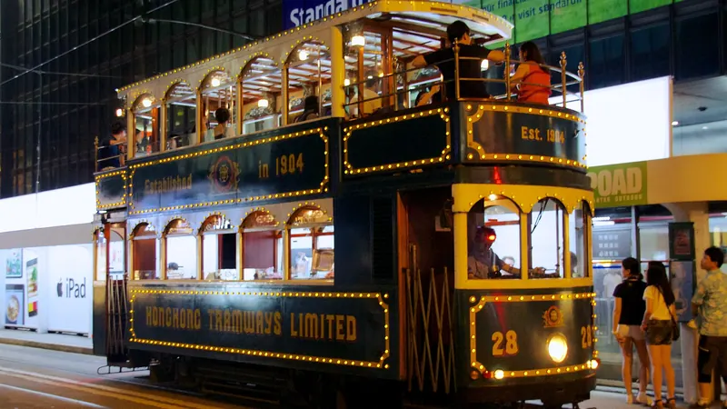 A dark green Hong Kong double-decker tram trimmed with lights at a stop at night, passengers on its open upper deck