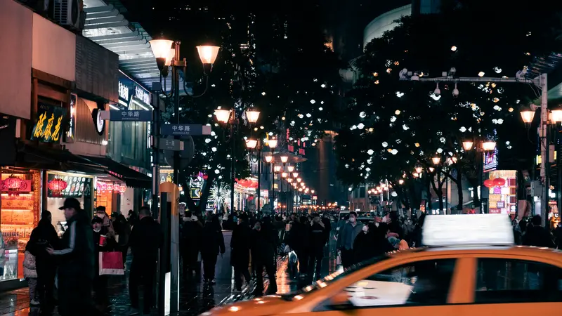 Una calle peatonal abarrotada de noche en una ciudad china, con farolas encendidas, letreros de tiendas y un taxi amarillo en primer plano
