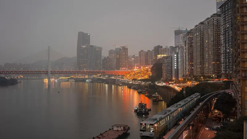 Chongqing at dusk under grey haze, with a monorail train running beside the river and lit towers and a bridge beyond
