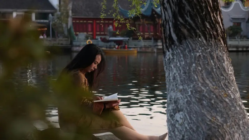 Mulher sentada à beira de um lago lendo um livro ao lado de um tronco de árvore, com construções tradicionais chinesas do outro lado da água, em Ningbo