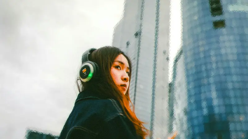 Young woman wearing large headphones looking over her shoulder in front of glass skyscrapers