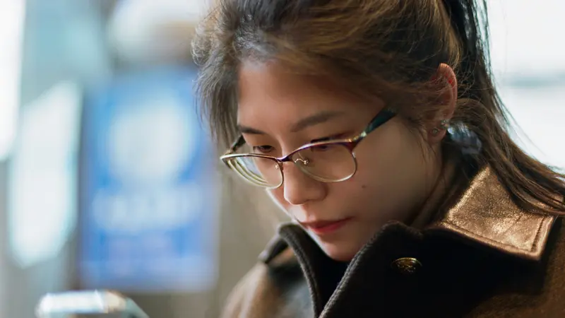 A young woman in glasses and a brown coat reading something on her phone