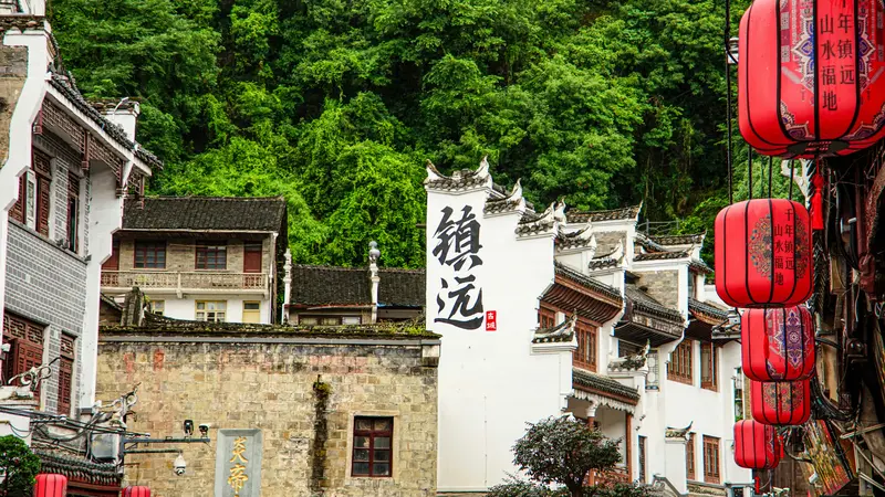 Construções antigas caiadas em Zhenyuan, na China, com os caracteres 镇远 pintados numa parede, lanternas vermelhas à direita e um morro coberto de mata ao fundo