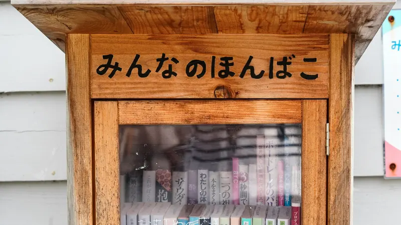 Pequena caixa de madeira para troca de livros, com porta de vidro e a inscrição em hiragana みんなのほんばこ, cheia de livros de bolso