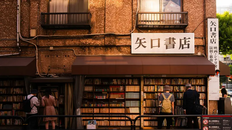 Shoppers browsing the outdoor shelves of a used bookstore on a street in Jimbocho, Tokyo, in warm evening light