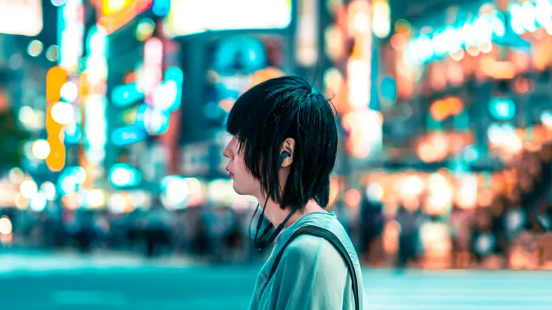 Person in a teal shirt wearing an earphone, standing in front of blurred neon signs on a Tokyo street at night