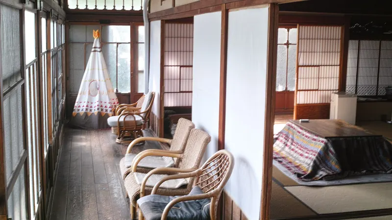 An old Japanese house with a wooden veranda of wicker chairs beside a tatami room with a quilted kotatsu table