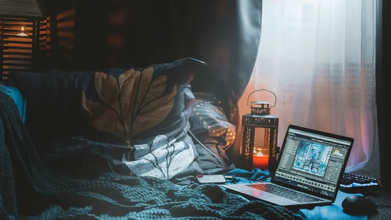 An open laptop on a bed beside a lit lantern and a window with sheer curtains in a dim bedroom
