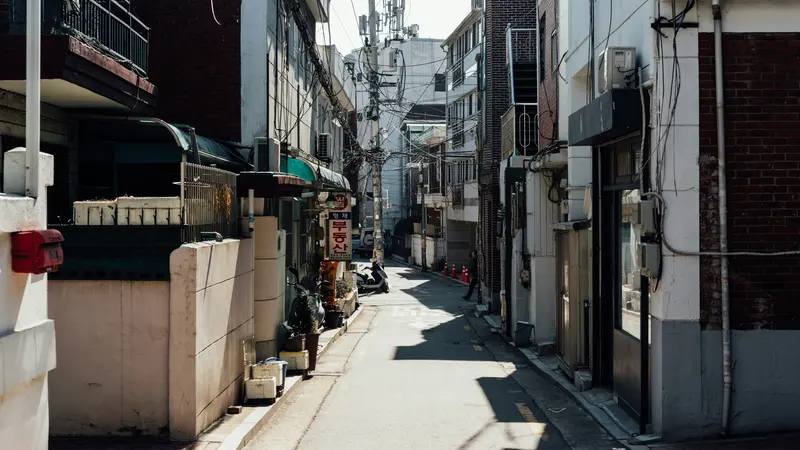 Un estrecho callejón residencial de Seúl bañado por el sol entre edificios bajos, con cables eléctricos en lo alto y algunas personas caminando a lo lejos
