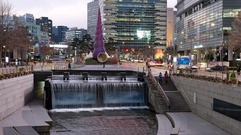The Cheonggyecheon stream walkway in central Seoul at dusk, with a small waterfall, steps and office towers