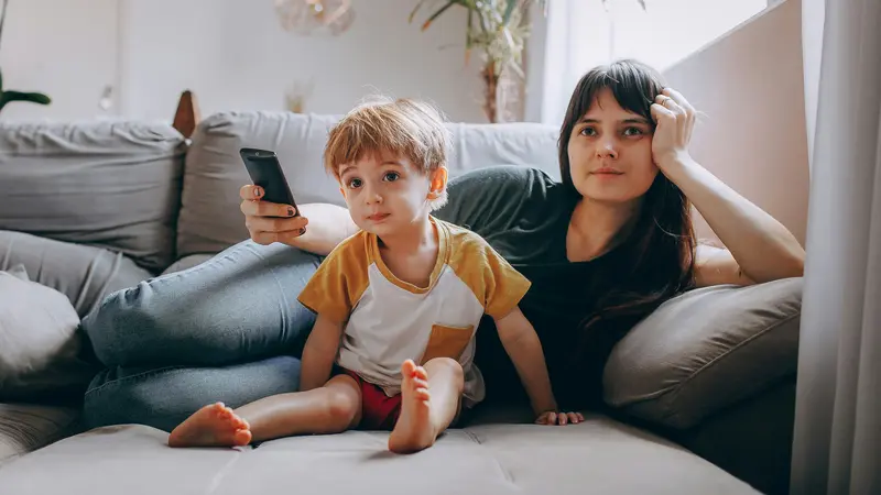 A young boy and a woman holding a remote control sitting on a gray sofa watching TV