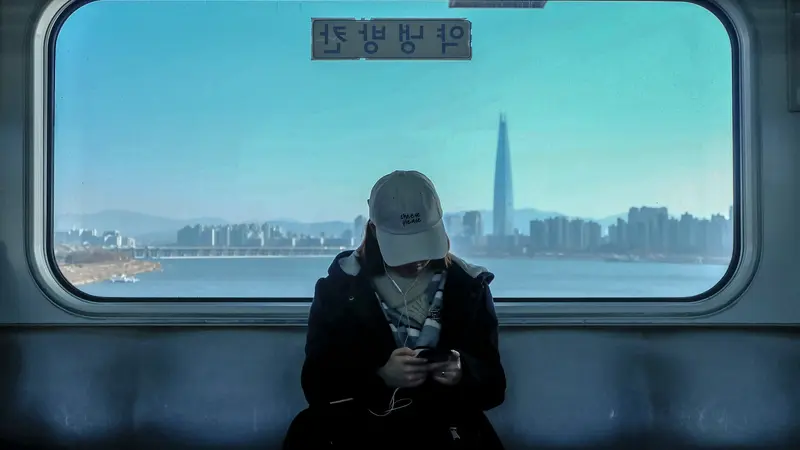 A woman in a cap and coat wearing earbuds on a Seoul train, with the Han River and city skyline in the window behind her