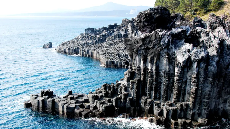 Dark basalt column cliffs of the Jeju Island coast meeting blue sea, with a mountain on the horizon