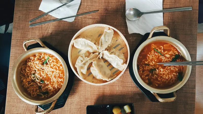 Two pots of instant ramyeon and a steamer basket of dumplings on a wooden table, seen from above, with spoons and chopsticks