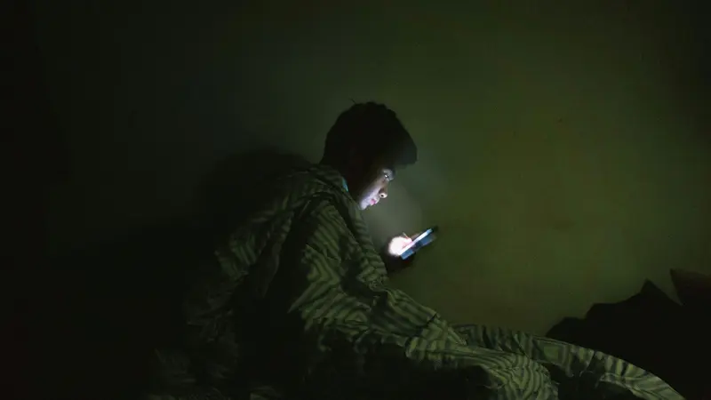 A man wrapped in a striped blanket looking at his phone in a dark room