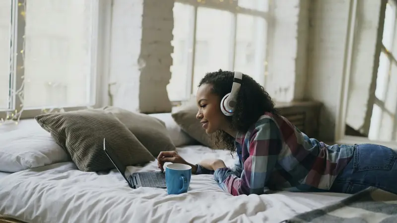 A young woman wearing headphones lying on a bed and watching a laptop, with a blue mug beside her