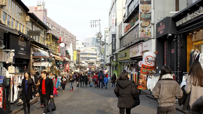 Shoppers walking down a busy street of clothing and snack shops in Hongdae, Seoul, on a sunny day