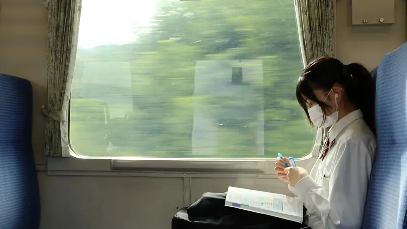 A student in a white shirt sitting by a train window with an open book on her lap, green scenery blurring outside