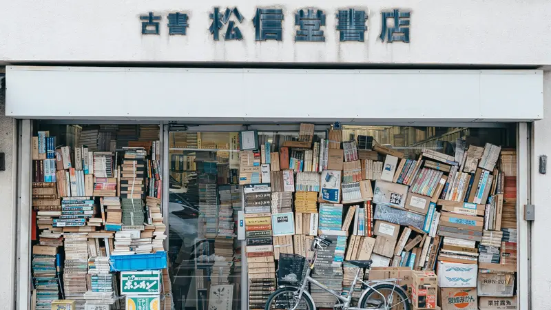 A used bookstore in Matsumoto, Japan, its front window piled high with stacked books and a bicycle parked outside
