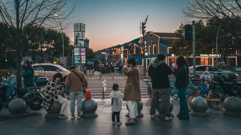 People and a small child waiting on a sidewalk by a crosswalk in Shanghai at dusk, with shared bikes and lit shopfronts behind them