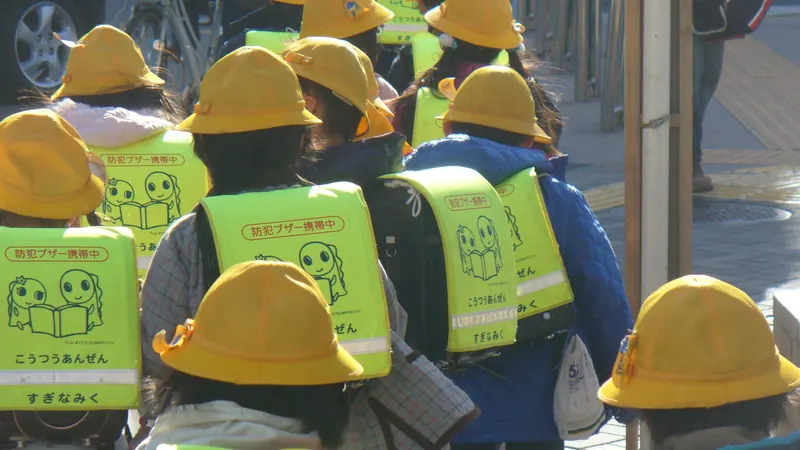 Japanese schoolchildren in yellow hats and safety vests over their randoseru backpacks, seen from behind on a walk