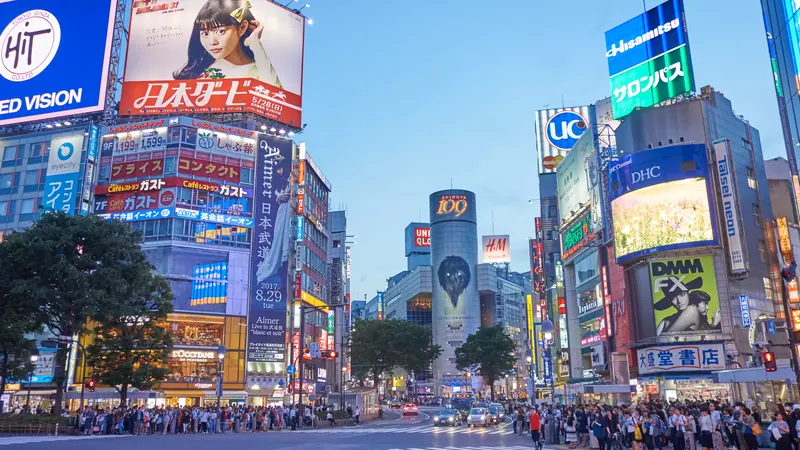 Crowds waiting at the Shibuya scramble crossing in Tokyo at dusk, under bright billboards and neon signs