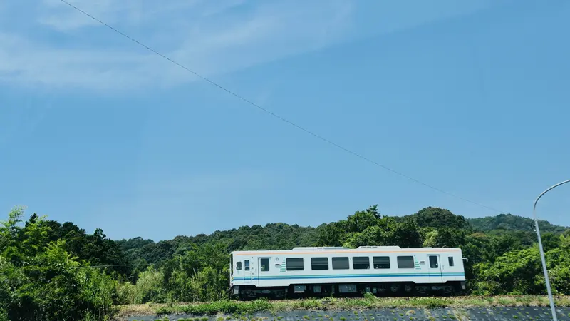 A single-car white train running along an embankment through green countryside under a clear blue sky in Aichi, Japan