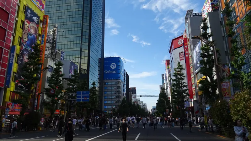Pedestrians walking down the car-free main street of Akihabara, Tokyo, between tall shop buildings under a blue sky