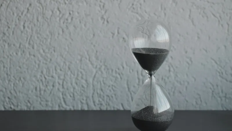 An hourglass with dark sand running, on a dark table against a textured wall