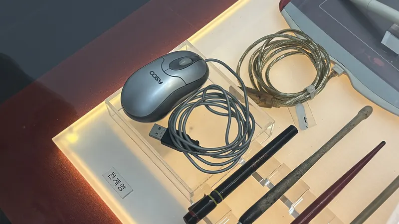 A computer mouse, drawing pen tablet and dip pens laid out on a lit display case at the Korea Manhwa Museum in Bucheon