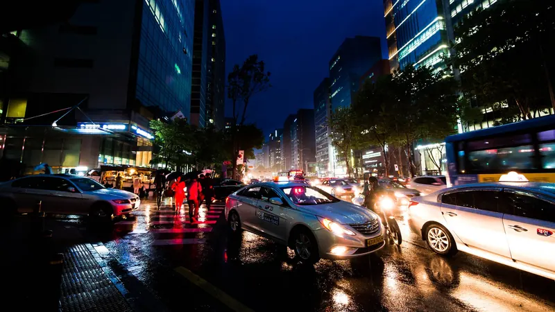 Taxis and cars with glowing headlights on a wet Seoul street on a rainy evening, between lit office towers