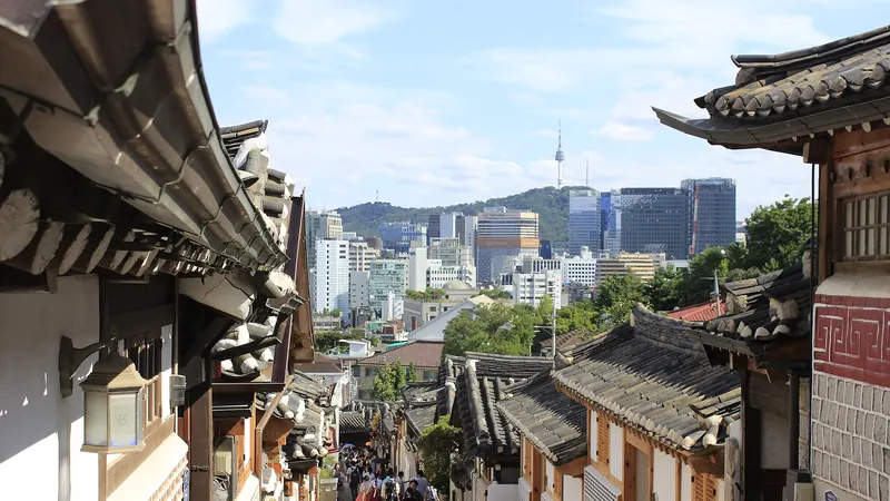 A sloping lane of tiled hanok roofs in Bukchon Hanok Village, Seoul, with N Seoul Tower and city towers in the distance