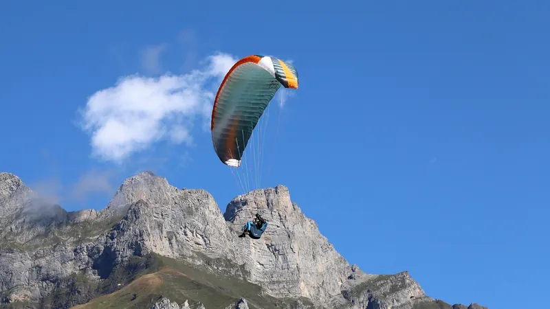 A paraglider with a green and orange wing soaring under a blue sky above jagged limestone peaks