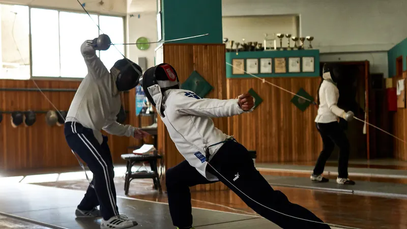 Two épée fencers in white jackets and masks lunging and parrying during a bout in a fencing club hall