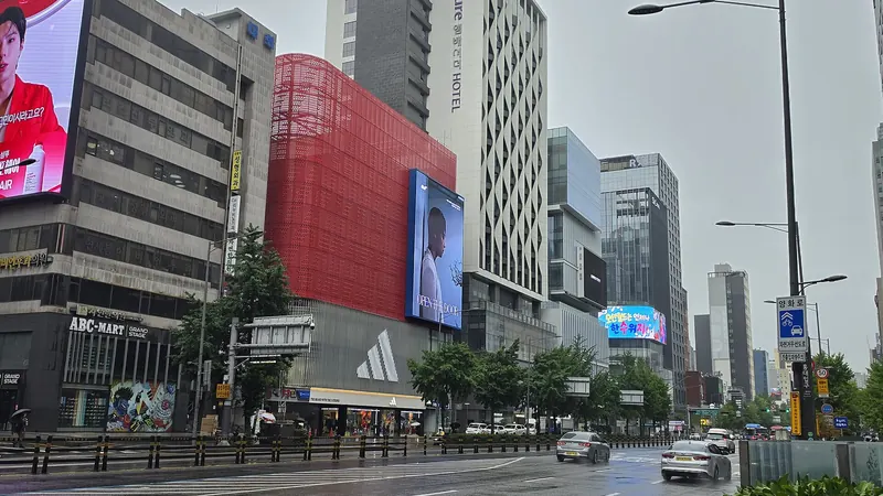 A rainy main road in Hongdae, Seoul, lined with buildings carrying giant video screens and billboards