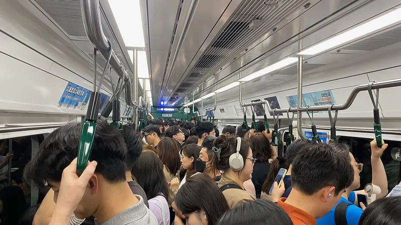 A packed Seoul subway car at rush hour, with commuters holding green straps and looking at their phones